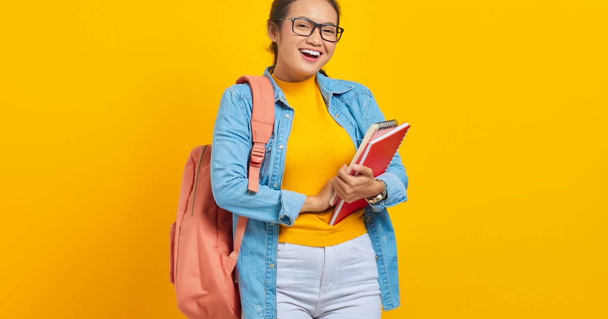 Portrait of cheerful young Asian woman student in casual clothes with backpack holding book and looking at camera isolated on yellow background