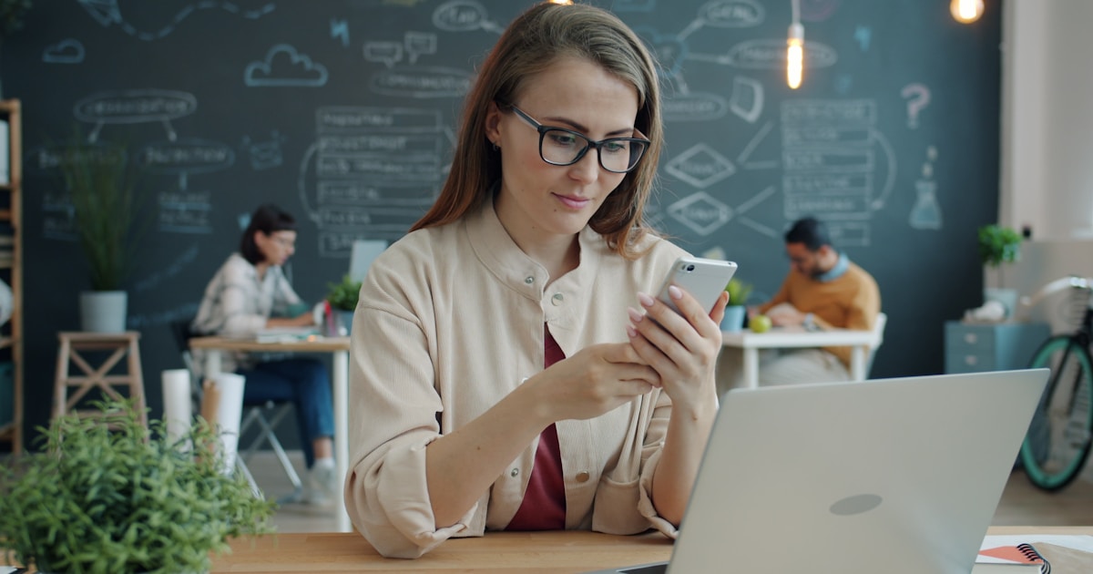 Young girl is using smartphone touching screen smiling sitting at desk in open space office room enjoying communication. People, workplace and devices concept.