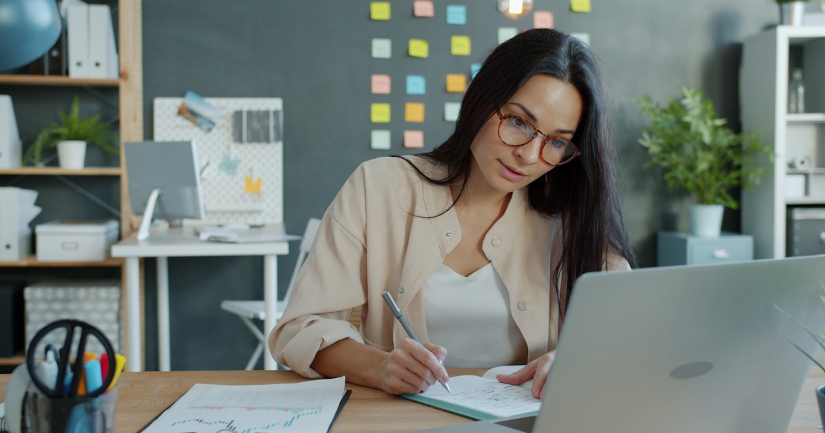 Young businesswoman in elegant clothing and glasses is writing in notebook and using computer smiling in office. Technology and occupation concept.